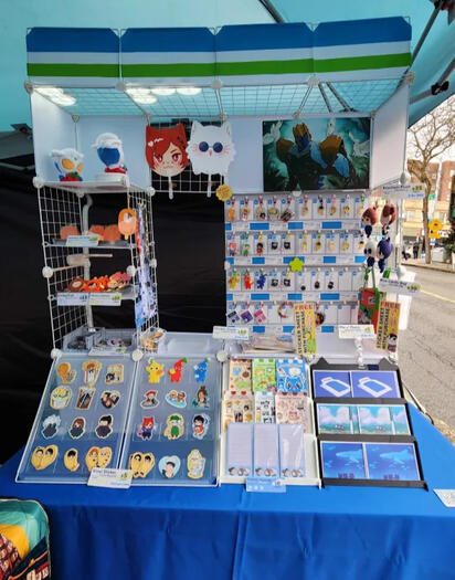 4ft artist alley table display for cid lny 2026 with stickers, charms, pvc fans, and prints of dunmeshi, jjk, ffxiv, original art