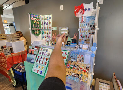 split artist alley table display for rinjin winter market 2024 with stickers, charms, pvc fans, and prints of dunmeshi, jjk, ffxiv, original art