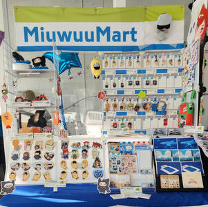 split artist alley table display for gojo hbd cupsleeve 2025 with stickers, charms, pvc fans, and prints of dunmeshi, jjk, ffxiv, original art